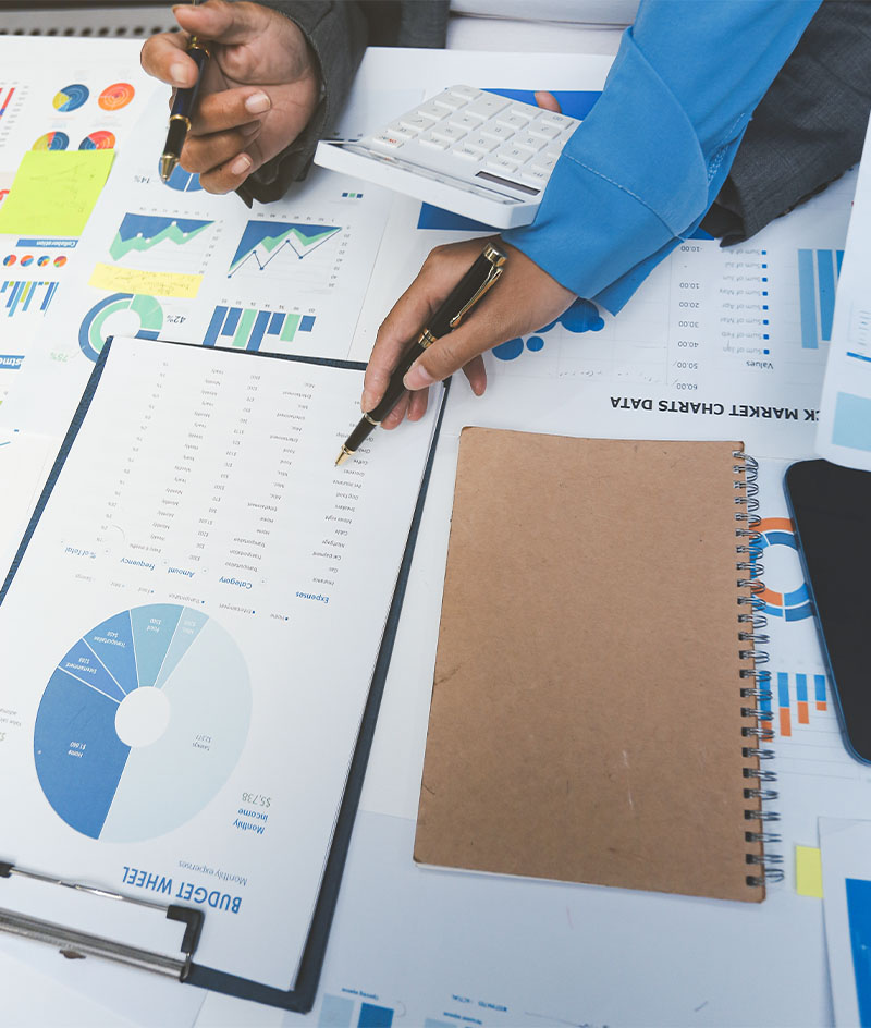 Two individuals sit at a desk covered with financial documents, charts, and graphs. One holds a pen and gestures toward a report on a clipboard, while the other uses a calculator. A spiral notebook lies on the desk alongside various printed charts showing data visualizations such as bar graphs, line graphs, and pie charts.
