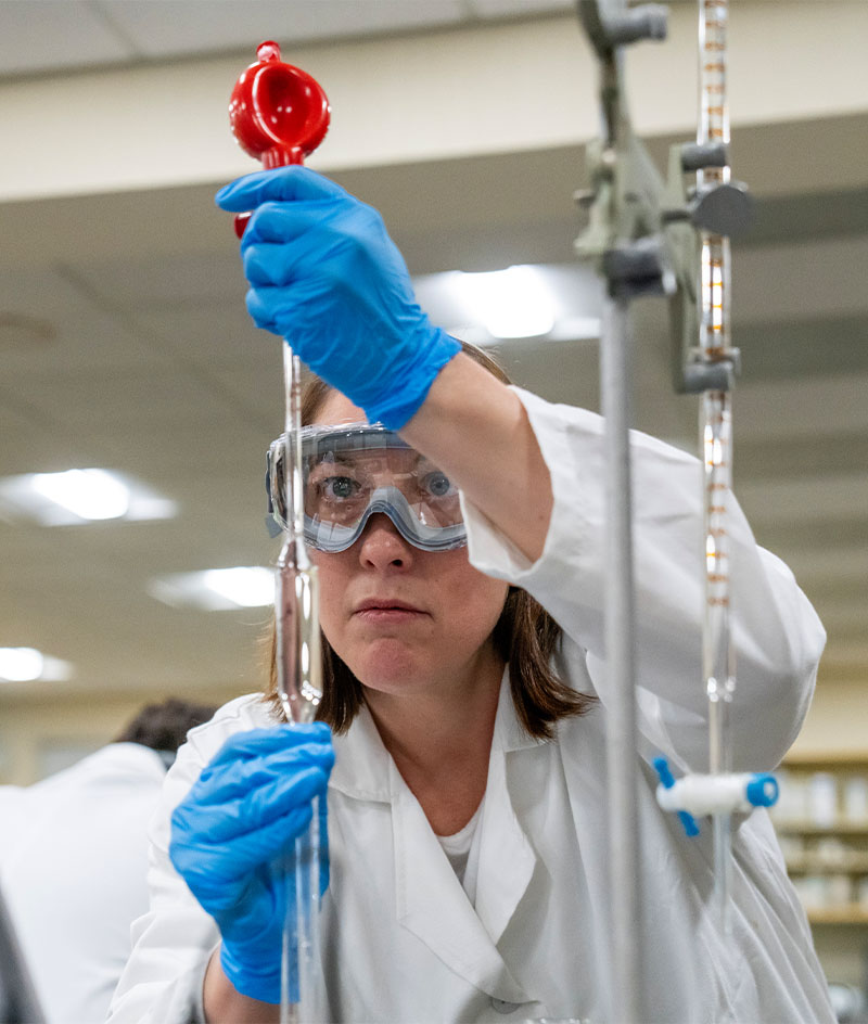A person wearing a white lab coat and blue protective gloves works with glass laboratory equipment, carefully adjusting a burette while holding a red funnel at the top. The individual is in a bright laboratory setting with shelves and scientific apparatus visible in the background.