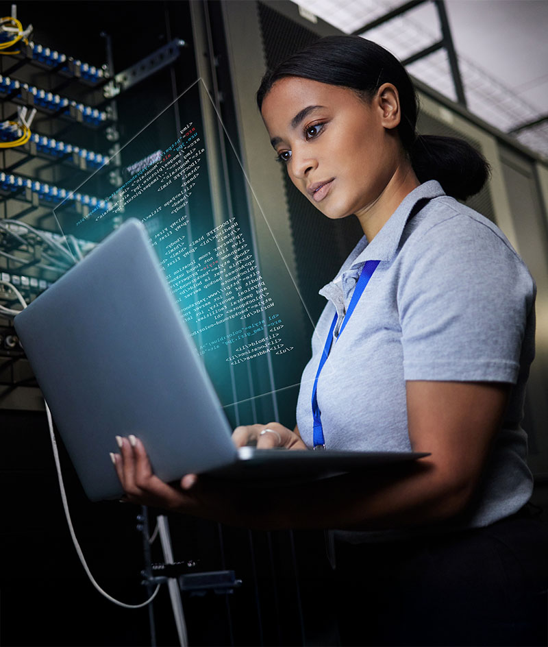 A person stands in a data center environment holding an open laptop while working beside tall server racks filled with networking equipment and cables. Digital code and interface graphics appear overlaid near the laptop screen, suggesting system monitoring, programming, or cybersecurity activity. The setting is dimly lit with focused lighting on the equipment, emphasizing a technical, high‑security workspace.