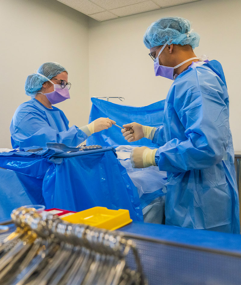 Two people wearing blue surgical gowns, masks, gloves, and hair covers work at a draped table inside a bright indoor room. Medical instruments are arranged in the foreground, with large windows and empty tables visible in the background.