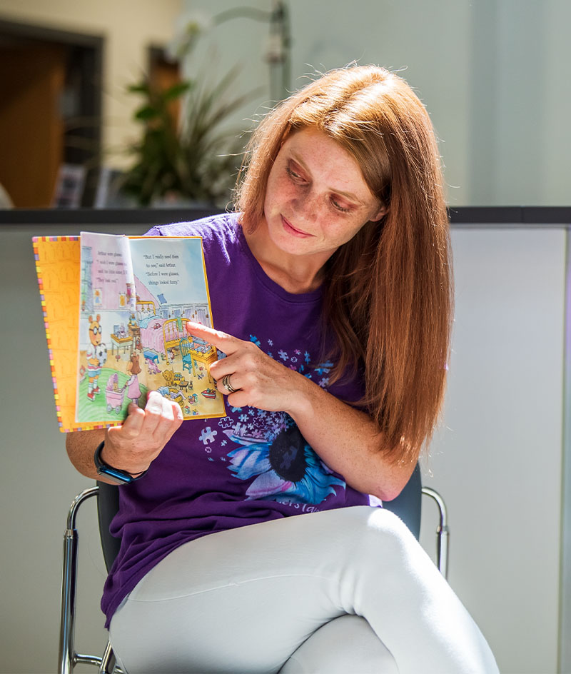 A person seated on a chair indoors reads from a colorful illustrated book, holding it open and pointing to a page. In the foreground is a display rack with children’s books, and in the background are office items such as a phone and stacked trays.