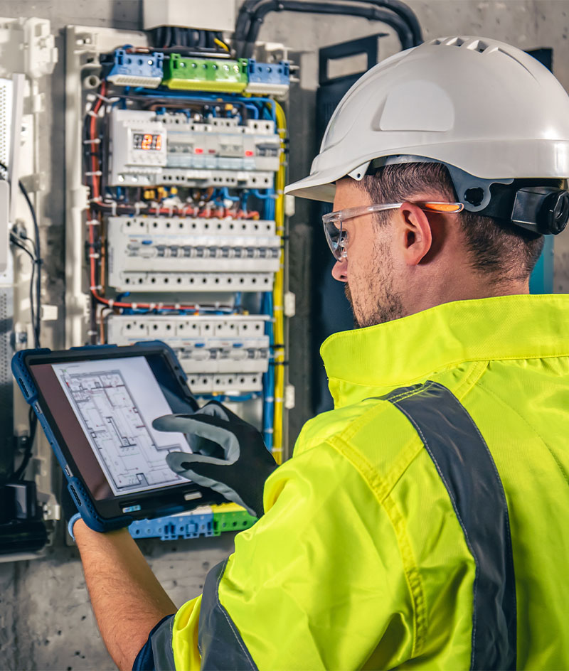A worker wearing a white hard hat, high‑visibility yellow jacket, gloves, and safety glasses stands in front of an open electrical control panel filled with circuit breakers, wiring, and components. The worker holds a rugged tablet displaying an electrical schematic, indicating inspection, troubleshooting, or maintenance work in an industrial or utility setting.
