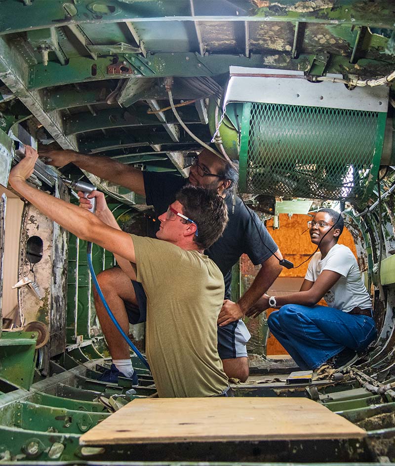 Interior of an aircraft fuselage under repair, showing three people working inside a narrow metal compartment with exposed ribs, wiring, and panels; one person uses a tool overhead while others assist nearby, with a wooden platform on the floor and green-painted structural components surrounding the workspace.