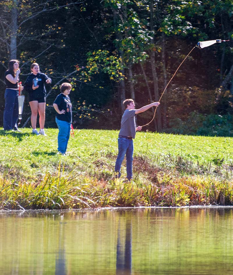 A group of people stands on a grassy bank beside a pond, with one person casting a long line or net over the water while others observe nearby; trees with dense foliage form the background, and the water reflects the green shoreline in bright daylight.