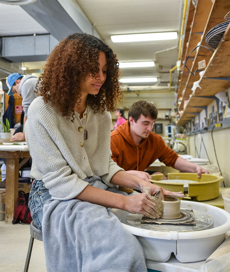 A person shapes clay on a pottery wheel in a studio classroom, hands coated with wet clay as a cylindrical vessel forms at the center of the spinning wheel; shelves, tools, and other people working at nearby wheels fill the well-lit workshop in the background.