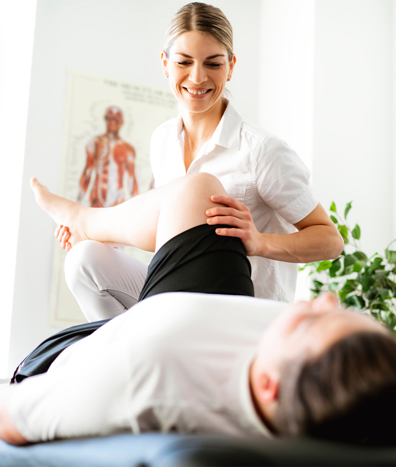 A clinician guides a patient through a leg stretch while the patient lies on a padded treatment table in a bright medical office. Anatomical charts and a skeleton model are visible in the background, along with a leafy plant near a window.