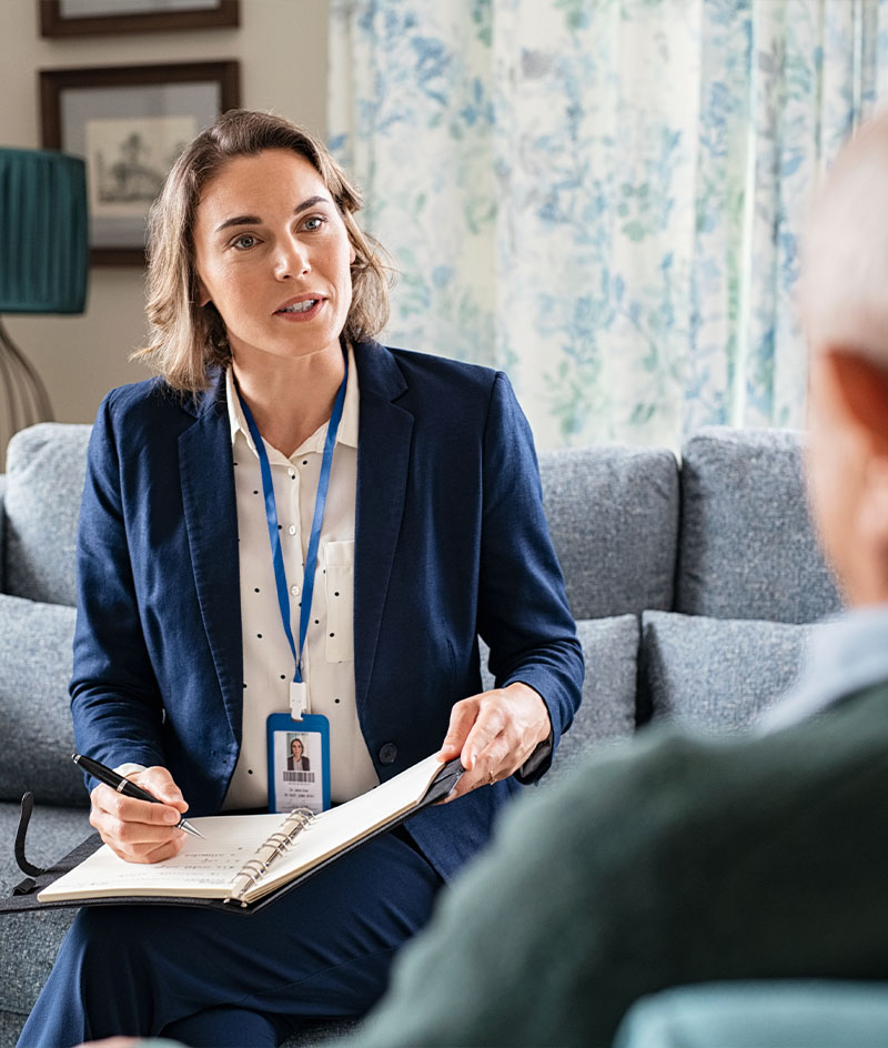 A person sits on a sofa in a softly lit living-room setting, holding a notebook and pen while taking notes during a conversation with another person seated nearby; framed artwork, patterned curtains, and upholstered furniture create a calm, professional indoor environment.