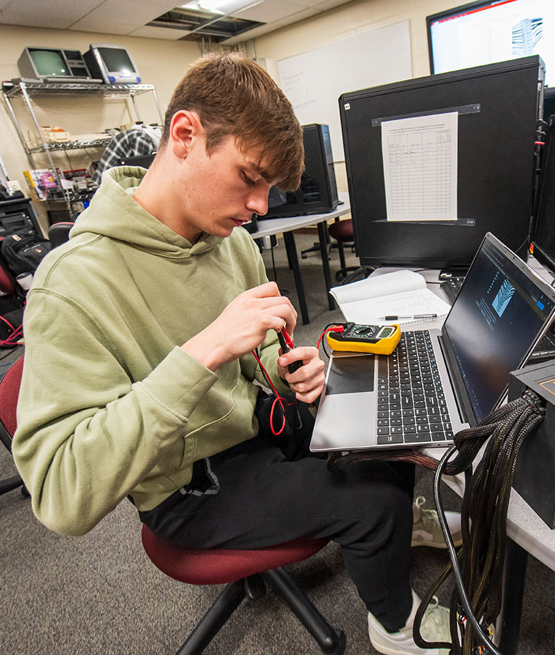 A person sits at a workstation in a computer lab, using a multimeter to test electronic components connected to a laptop; cables, a notebook, and multiple computer monitors are arranged on desks in a classroom-style technical environment.