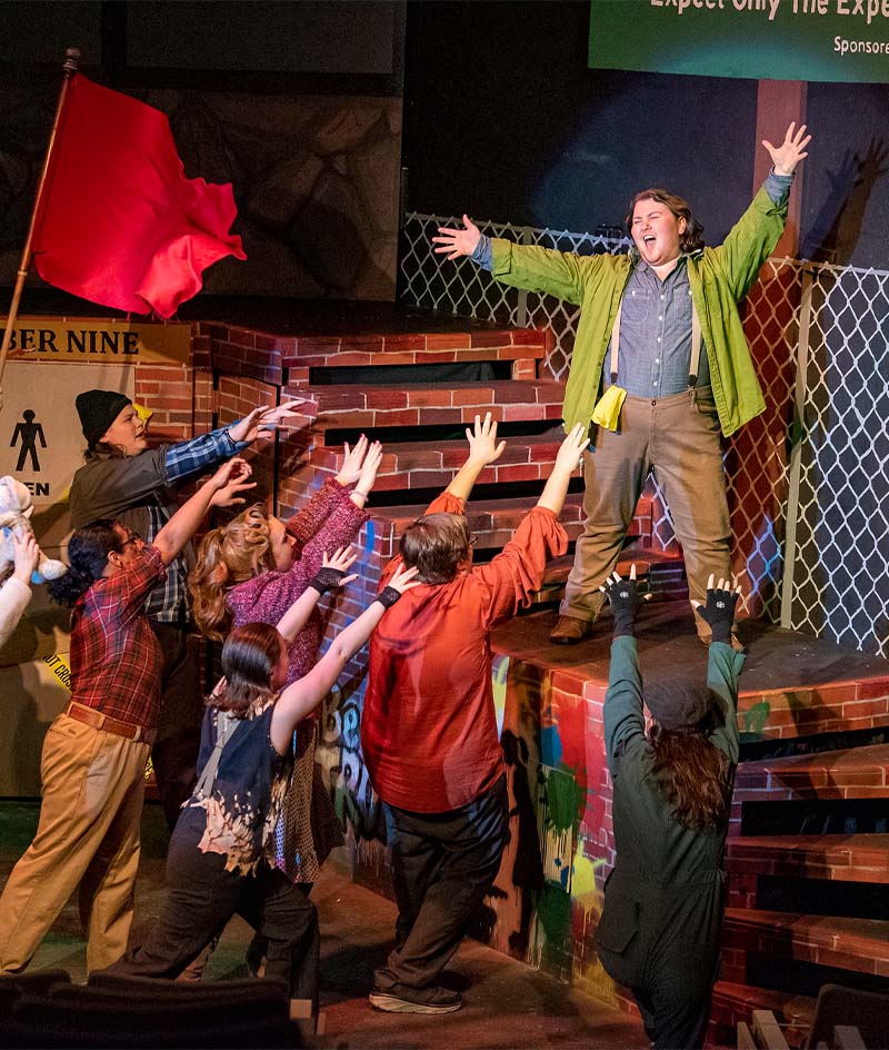 A stage performance shows several people reaching upward toward another person standing on stacked brick steps with arms raised. The set features brick walls, a chain-link fence, and protest-style signs, with dramatic stage lighting illuminating the ensemble scene.
