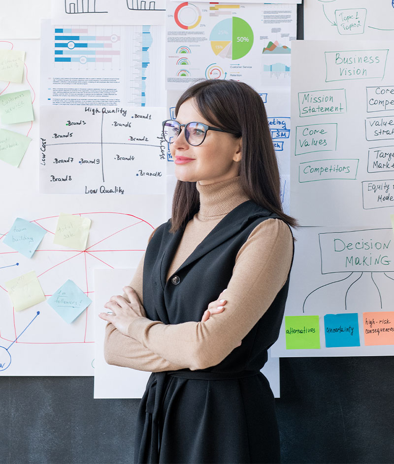 A person stands with arms crossed in front of a dark wall covered with pinned sheets of paper showing charts, graphs, diagrams, handwritten notes, and colorful sticky notes. The setting resembles a workspace or planning area focused on data analysis, strategy, or problem-solving, with papers arranged densely across the wall.