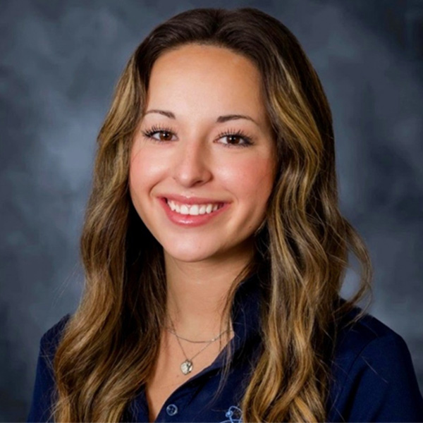 A person with long wavy hair featuring light and dark highlights is shown against a white background. The individual is wearing a navy collared shirt and layered necklaces, including a small pendant.