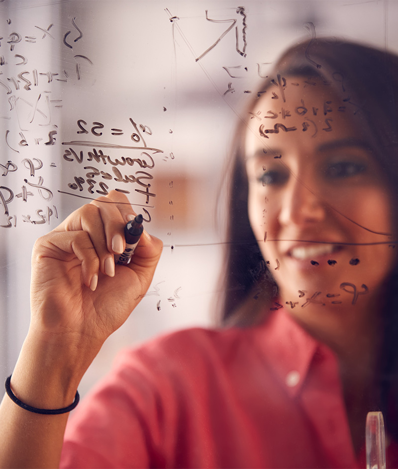 A person writes mathematical equations and symbols with a marker on a transparent board, viewed close up from the side, with additional handwritten formulas visible in the background.