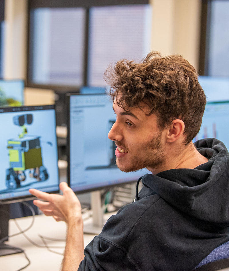 A student sits at a computer workstation in a lab, gesturing toward multiple monitors displaying 3D CAD models of mechanical components, with additional computer stations visible in the background.