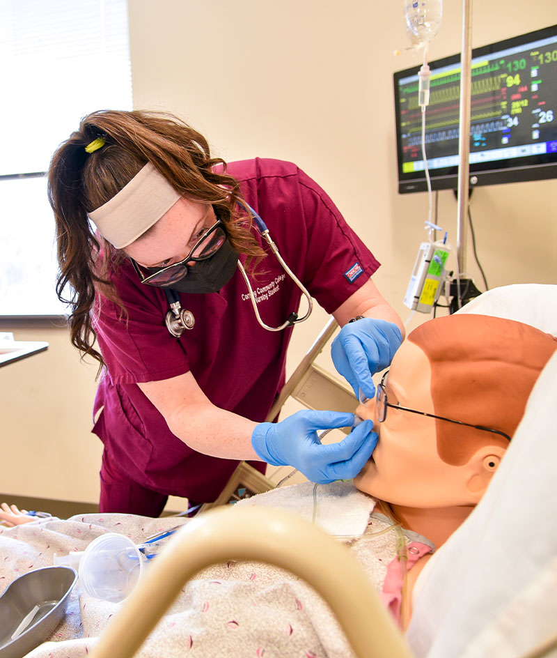 Nursing student practices patient care skills on a medical simulation mannequin in a clinical lab, using monitoring equipment and hands‑on techniques.